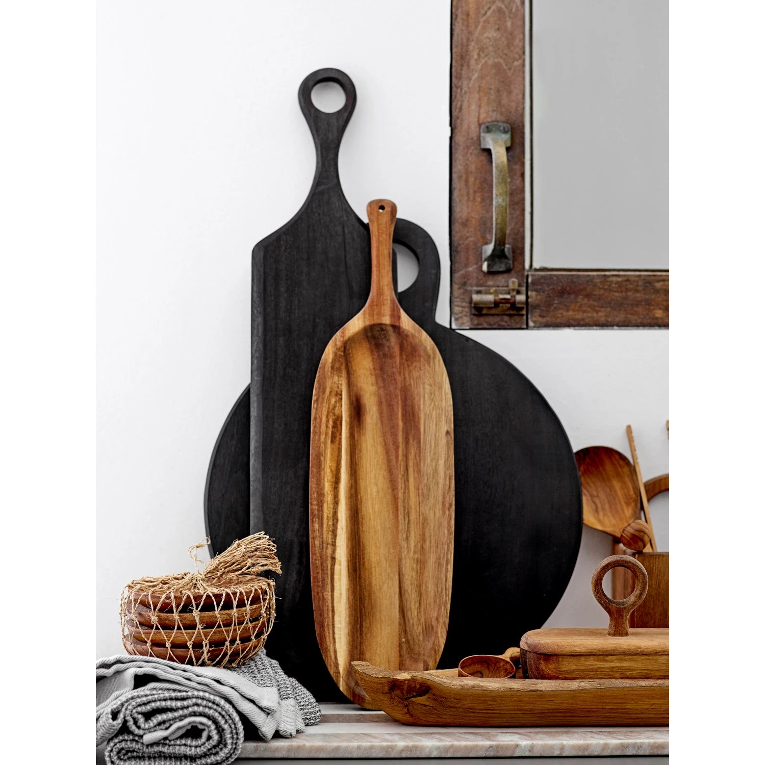 beautiful black acacia wood cutting board displayed upright in a kitchen with other warm wood boards and trays, and soft dish towels.