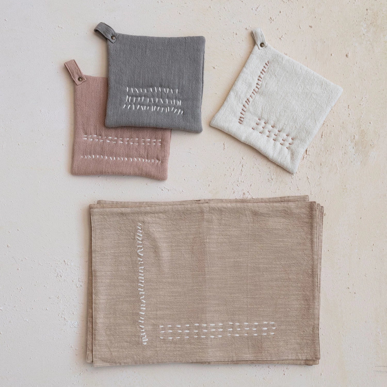 Embroidered cotton slub pot holders in grey, linen, and washed terracotta. Pictured next to embroidered beige kitchen towels