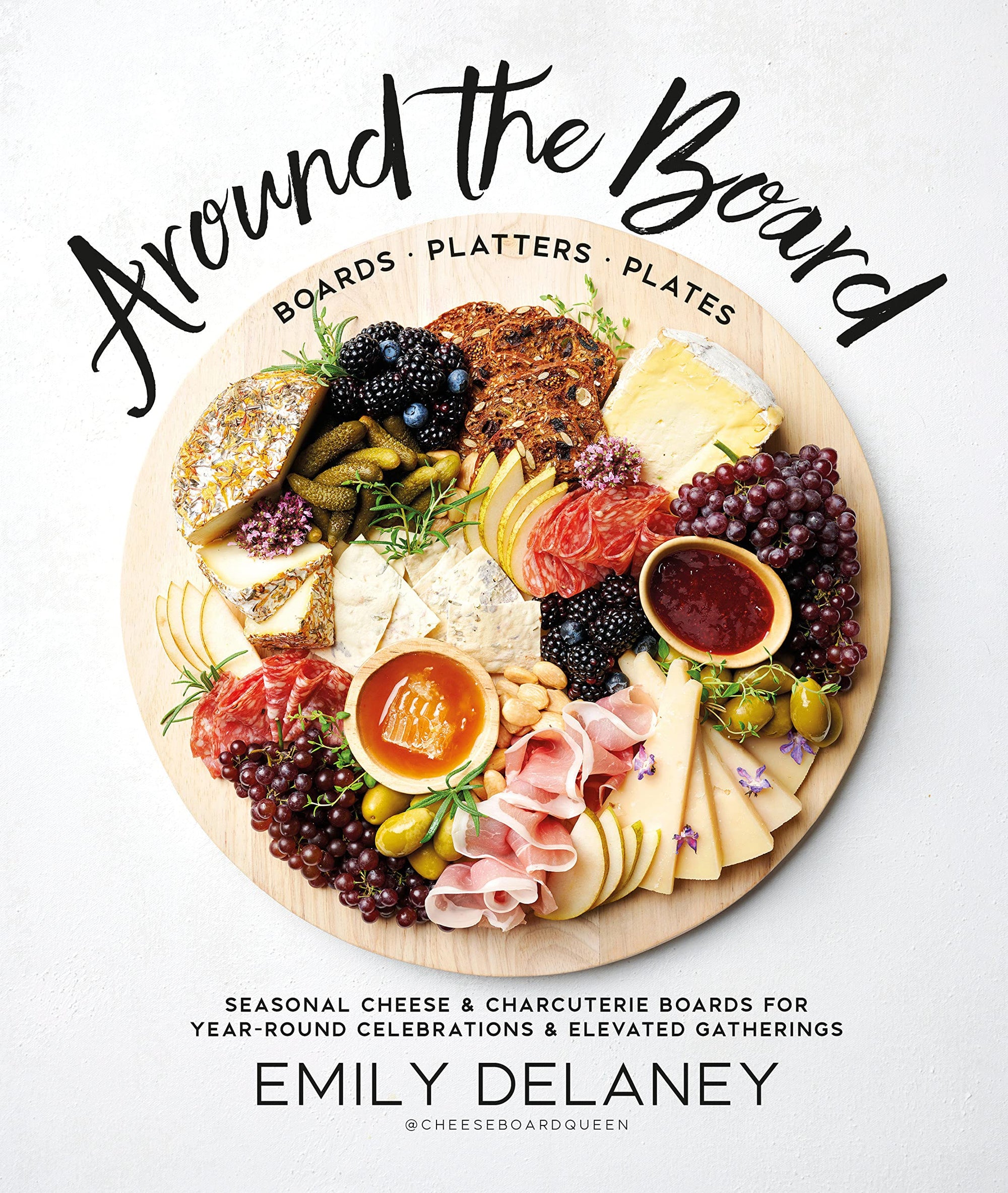 Front cover of charcuterie cookbook "Around the Board". Displayed on a rustic natural cutting board is a variation of cheeses, fruits, nuts, and jams in a coordinating display. Modern charcuterie display. Charcuterie gatherings. 