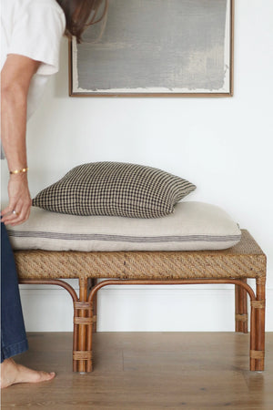 A woven rattan bench with a light beige cushion and a black-and-beige busy windowpane throw pillow, shown in a minimalist room with a person partially visible on the left.