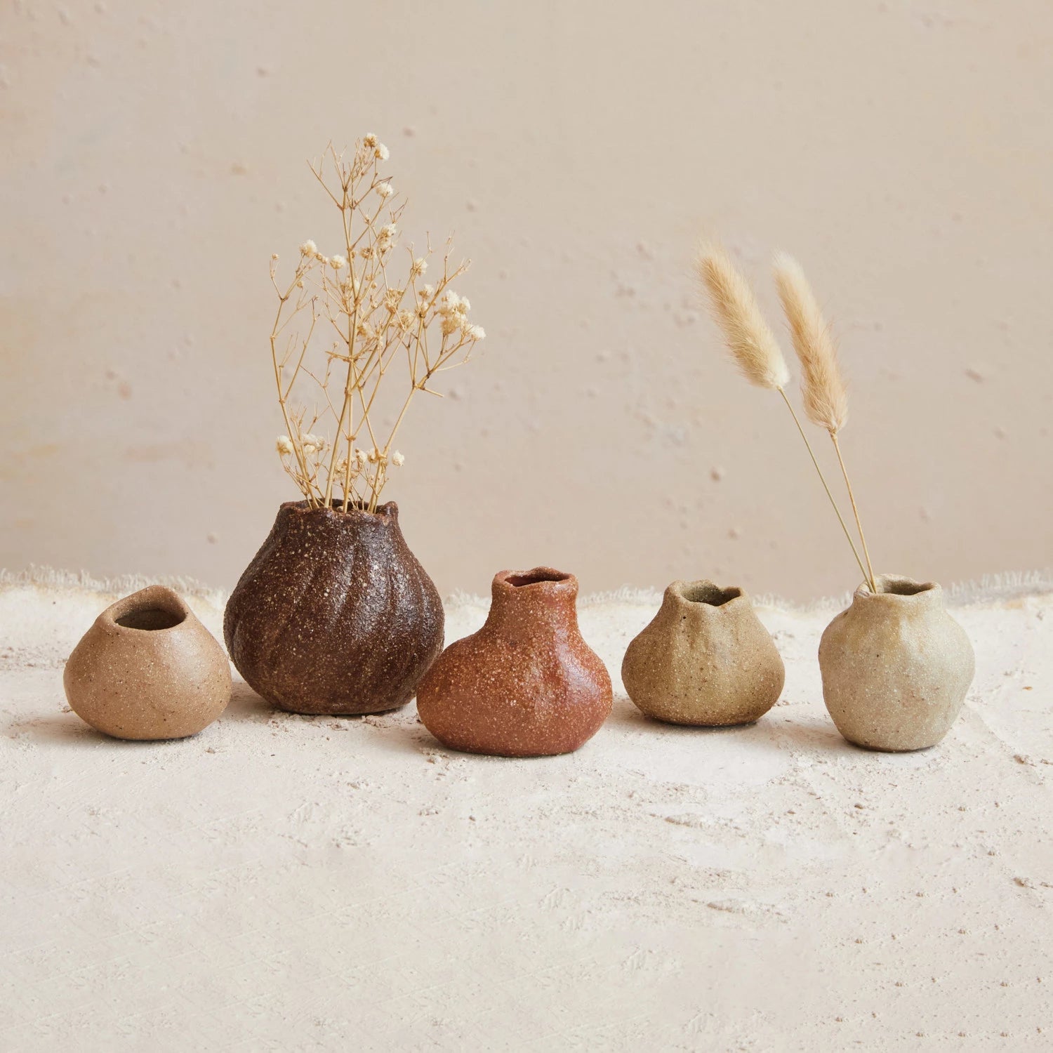 Collection  of organic shaped stoneware two inch vases in varying colors and textures displayed with dried floral stems. 