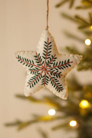 Cream colored felt star ornament embroidered with green and brown detailing hanging from a tree with a blurred background. 