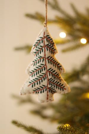 Cream colored felt Christmas tree ornament embroidered with green and brown detailing hanging from a tree with a blurred background. 