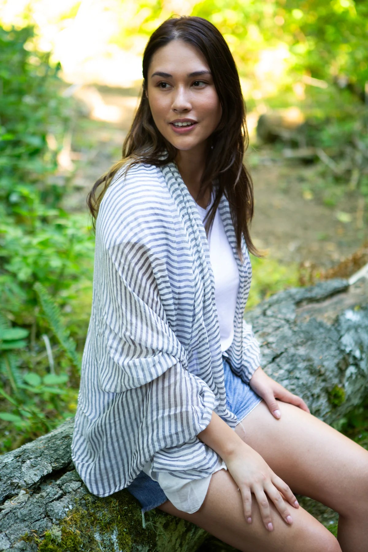 A woman sitting outdoors on a mossy log, wearing a white and black striped scarf over a white top and denim shorts.