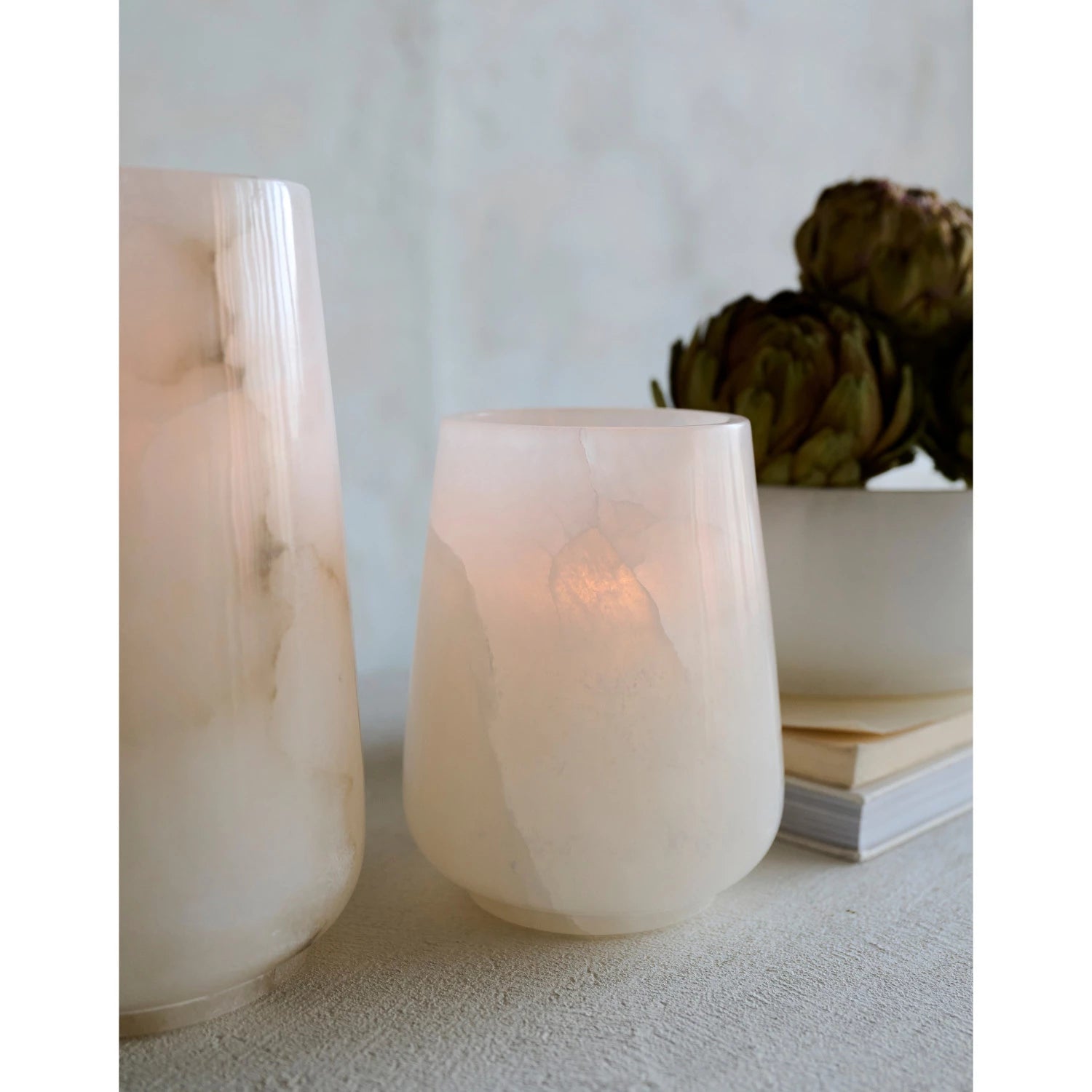 Two alabaster marble hurricane candle holders on textured neutral surface with a blurred background. 