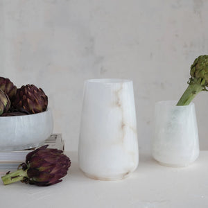 Large and small alabaster marble hurricane candle votives on a light surface with green and purple artichokes around them.
