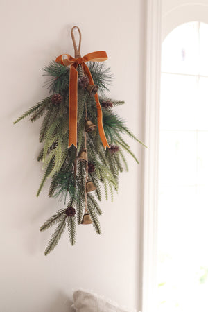 Decorative wall hanging garland with pine cones, bells, and rust brown velvet ribbon bow hanging from  a white wall.