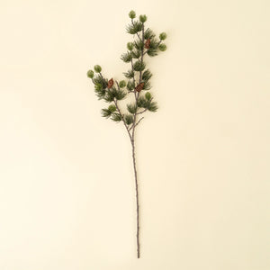 An artificial pine branch with green needles, small pinecones, and a long brown stem, displayed against a solid cream-colored background.
