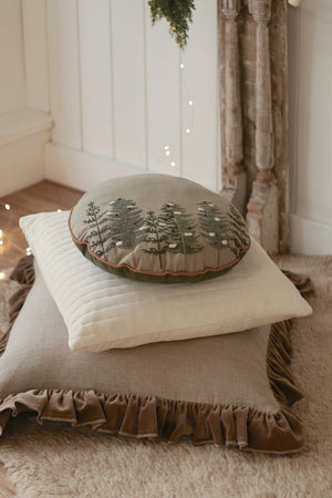 Stack of decorative pillows with a round embroidered holiday Alpine forest pillow on a soft rug surface.