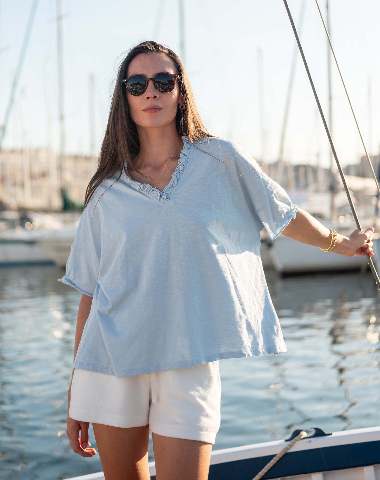 Woman in a light blue top and white shorts standing on a boat with sailboats in the background.