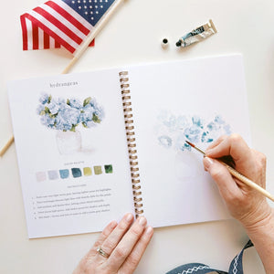 Person painting flowers in a sketchbook with an American flag and art supplies in the background