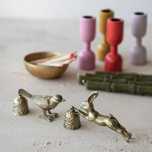 Brass animal figurine of bird and rabbit candle snuffers on a textured surface with colorful vases in the background. 