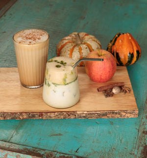 Two frothy drinks arranged on a wooden board with autumn props, including a striped glass of tan beverage topped with foam and a white drink with a metal straw, set on a turquoise wooden surface with pumpkins, an apple, and spices. Portland Syrups. 