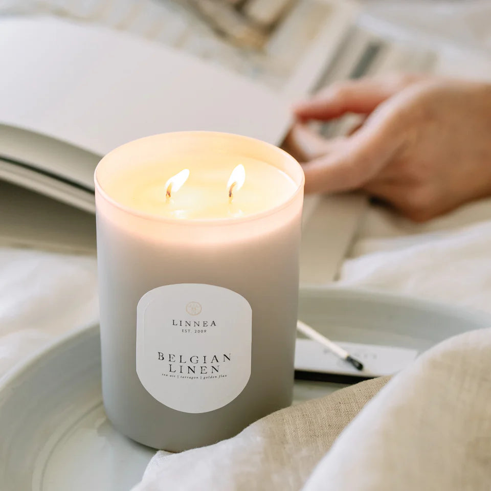 Candle labeled 'LINNEA Belgian Linen' on a surface with a blurred background of someone reading.