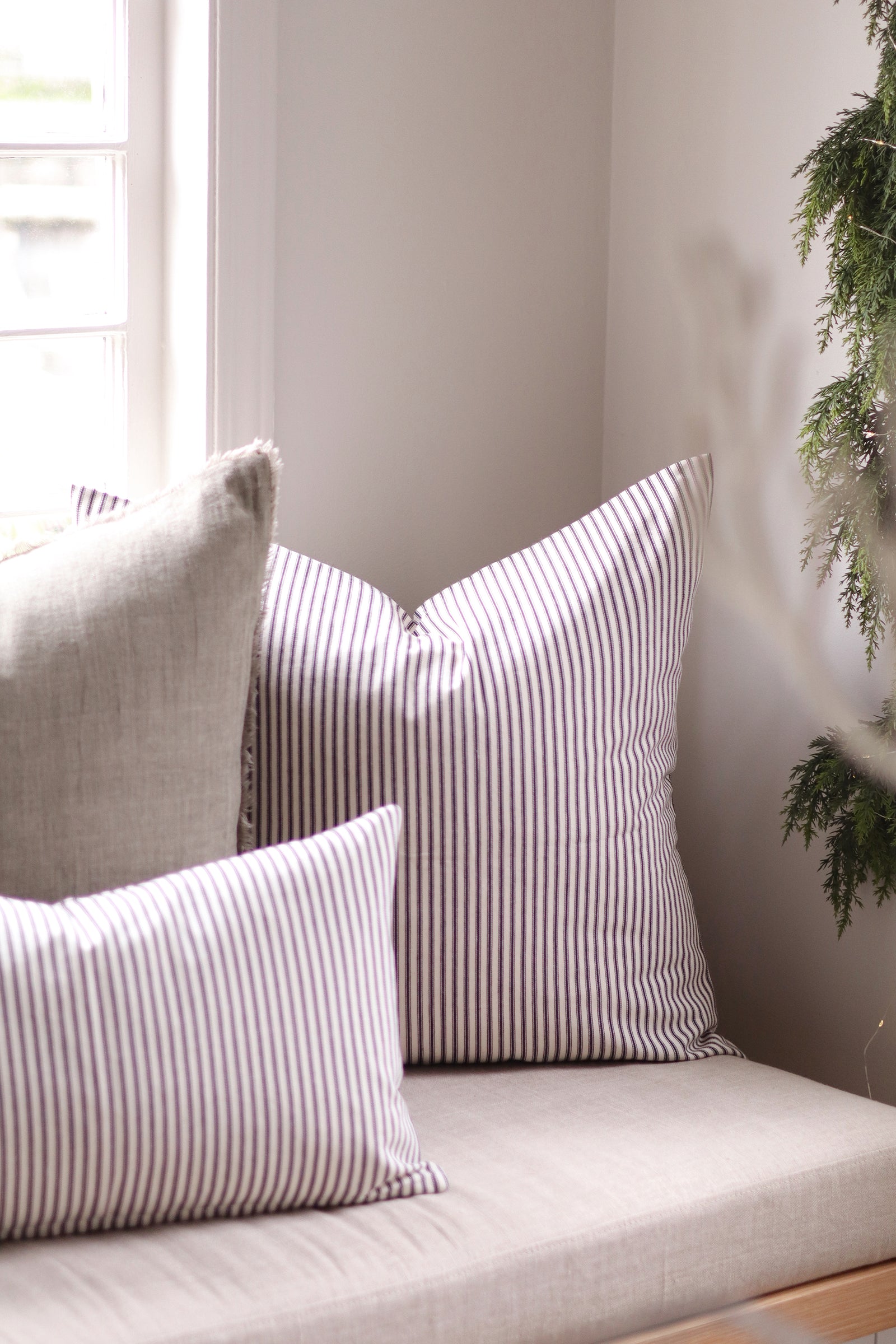 Three decorative throw pillows arranged on a light beige upholstered bench in a softly lit interior corner. Two pillows have narrow ticking vertical stripes in black and white, and one larger pillow is solid light beige.