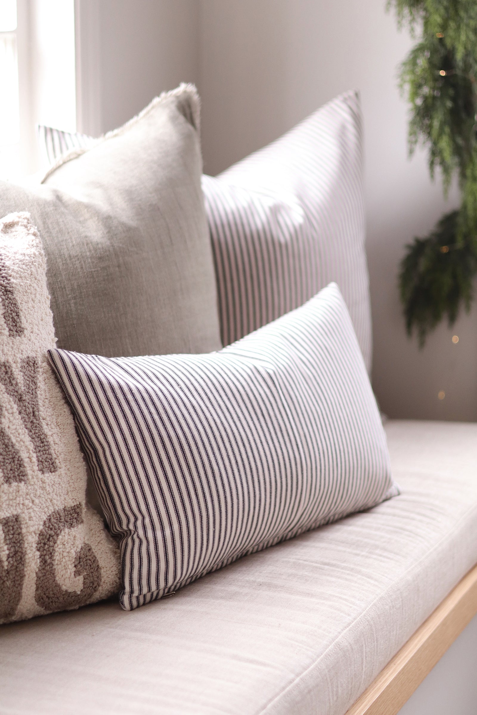 A cozy collection of neutral-toned throw pillows arranged on a light beige upholstered bench or window seat. The pillows include solid light gray, vertically striped black-and-white, and a textured cream pillow with taupe lettering, set against a softly lit interior background with a green plant visible on the right.