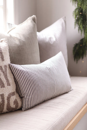 A cozy collection of neutral-toned throw pillows arranged on a light beige upholstered bench or window seat. The pillows include solid light gray, vertically striped black-and-white, and a textured cream pillow with taupe lettering, set against a softly lit interior background with a green plant visible on the right.