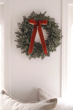 A green boxwood wreath with a large rust-brown velvet bow, hanging on a white wall above a light-colored sofa.