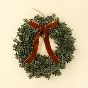 A green boxwood wreath with a large rust-brown velvet ribbon bow, hanging against a solid cream background.