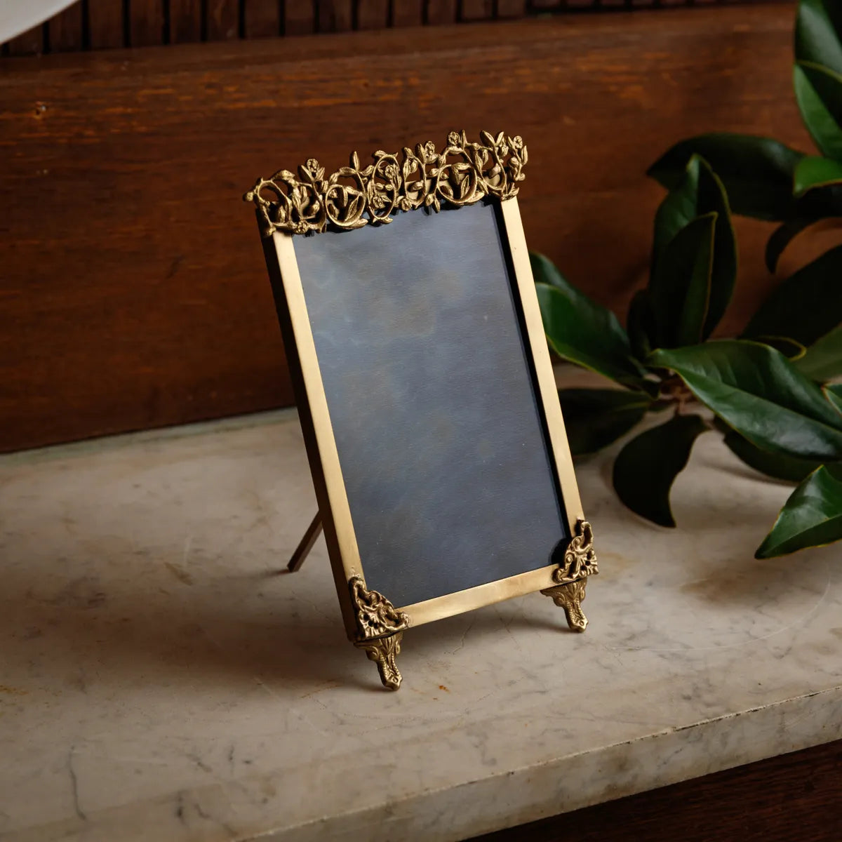 Art deco inspired vintage brass frame easel with floral motifs on a marble surface with a plant in the background. 