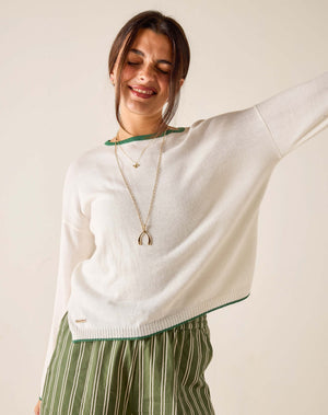 Woman wearing a white sweater with green trim and striped pants, standing against a plain background.