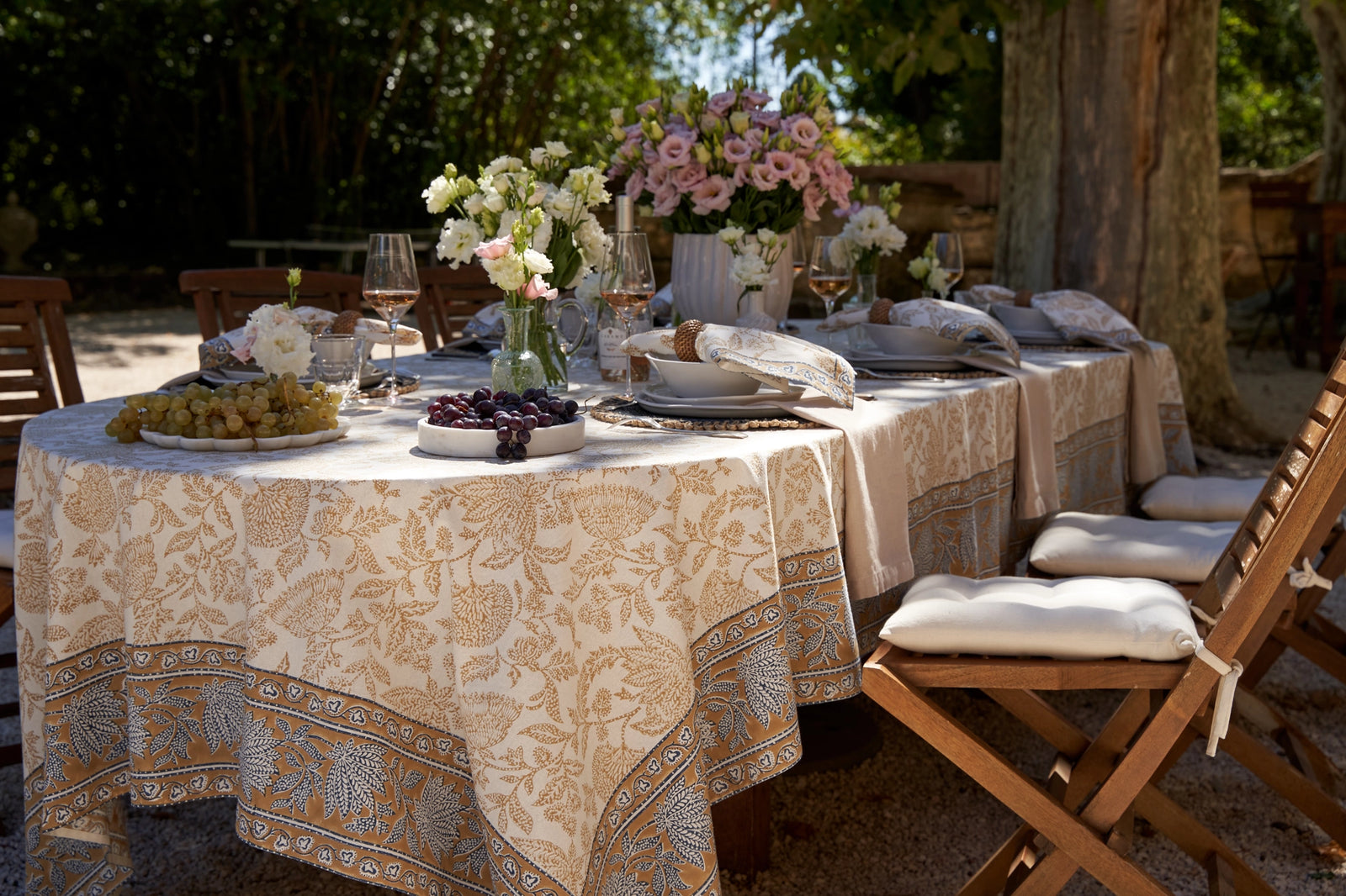 Outdoor dining table set with floral arrangements and place settings, surrounded by chairs. Blue and gold hued floral cotton tablecloth draped over long oval dining table. 