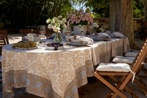 Outdoor dining table set with floral arrangements and place settings, surrounded by chairs. Blue and gold hued floral cotton tablecloth draped over long oval dining table. 
