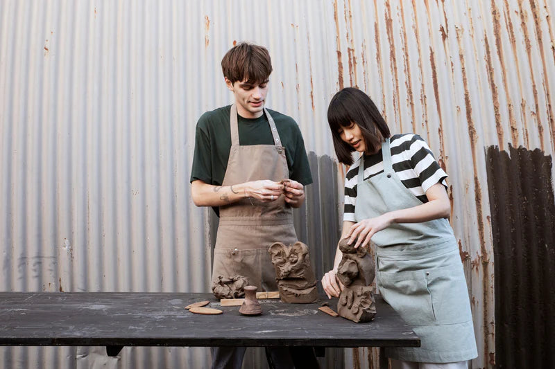 Two people working with clay sculptures in a workshop setting wearing organic cotton full sized aprons. The Organic Company. 
