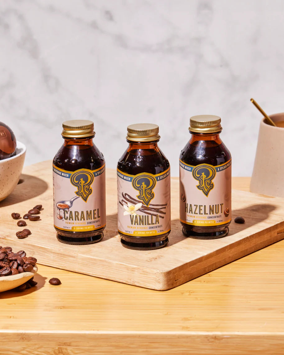 Three bottles of caramel, vanilla, and hazelnut syrup on a wooden tray with coffee beans and a cup in the background.