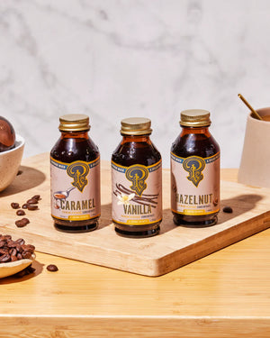 Three bottles of caramel, vanilla, and hazelnut syrup on a wooden tray with coffee beans and a cup in the background.