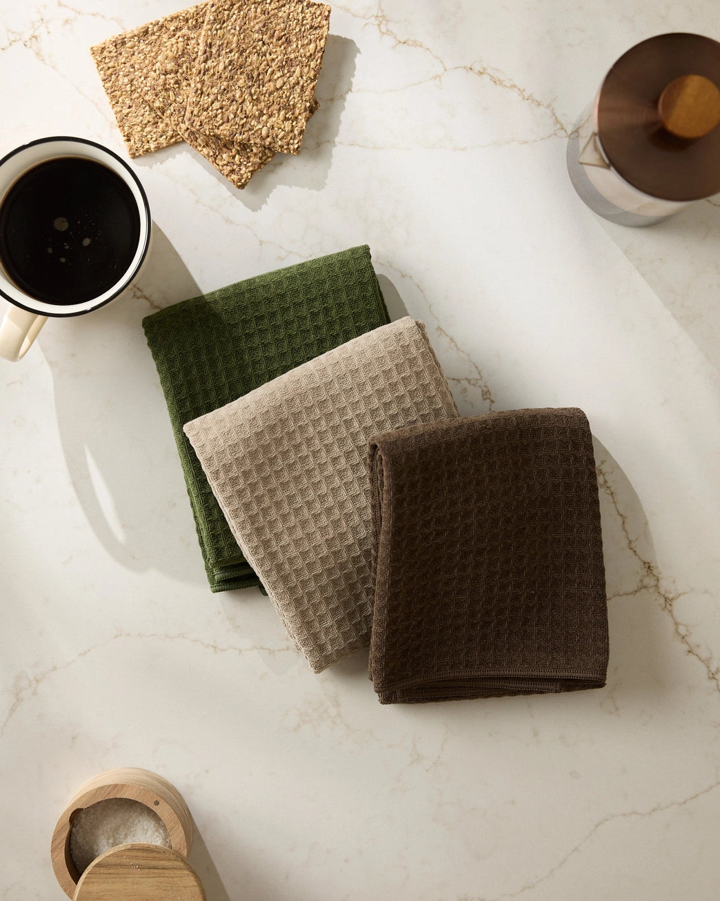 Set of three waffle-textured hand towels in green, beige, and brown on a marble surface with a cup of coffee.