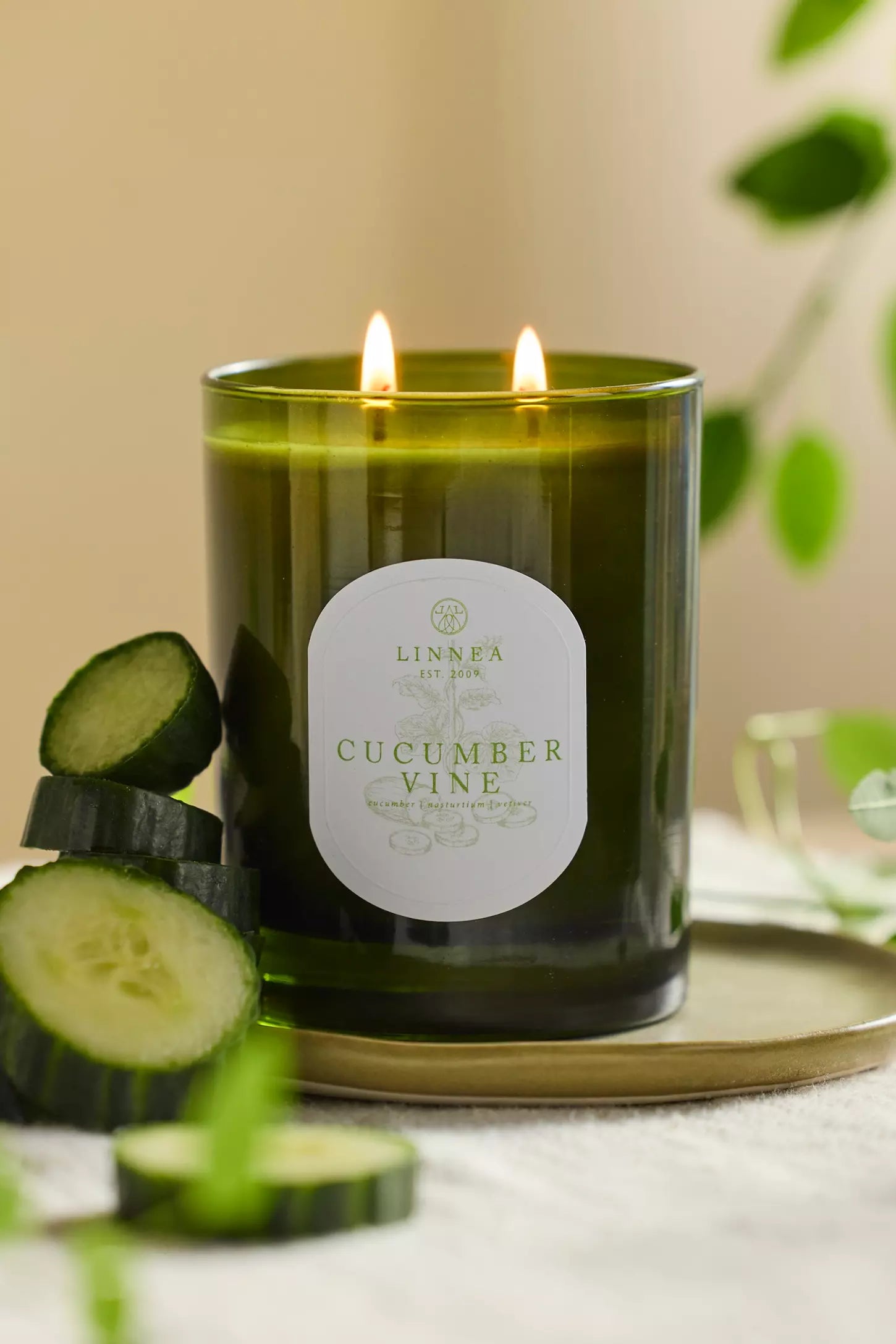 Candle labeled 'Cucumber Vine' with cucumbers stacked on a gold metallic tray against a neutral background. Linnea Botnik collection. 