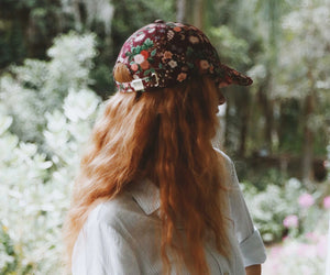 Person with long red hair wearing a dahlia floral baseball cap in a garden setting.