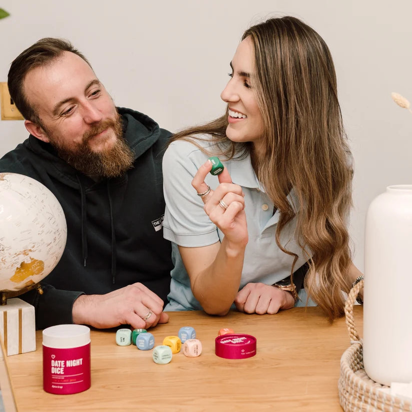 A man and a woman sit at a wooden table indoors, smiling and interacting with small colorful dice. A red tin labeled "DATE NIGHT DICE" is visible on the table, along with more dice and decorative objects like a globe and a white vase.