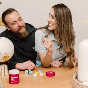 A man and a woman sit at a wooden table indoors, smiling and interacting with small colorful dice. A red tin labeled "DATE NIGHT DICE" is visible on the table, along with more dice and decorative objects like a globe and a white vase.
