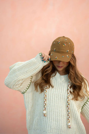 Woman wearing a beige knitted sweater and a brown ball cap with embroidered dahlias on it against a pink background. Rifle Paper Co. 