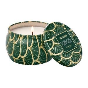 Green and gold patterned tin candle in Voluspa's scent Noble Fir Garland with a lit wick on a white background. 