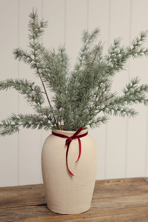 A rustic cream-colored ceramic vase with a textured finish, filled with green pine branches and tied with a red ribbon bow, set on a wooden surface against a white paneled wall.