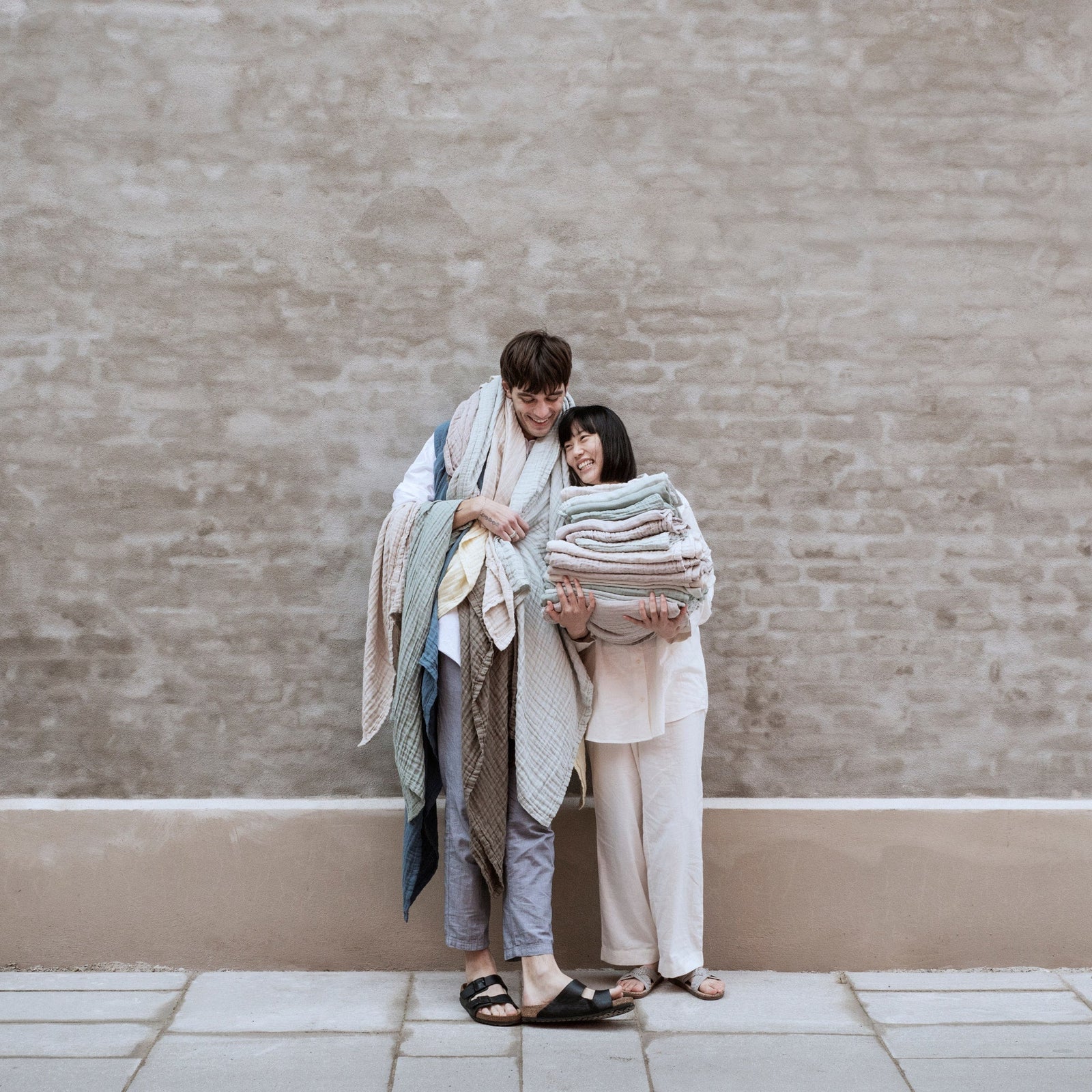 Two people holding stacks of folded organic cotton gauze bath towels against a beige brick wall.