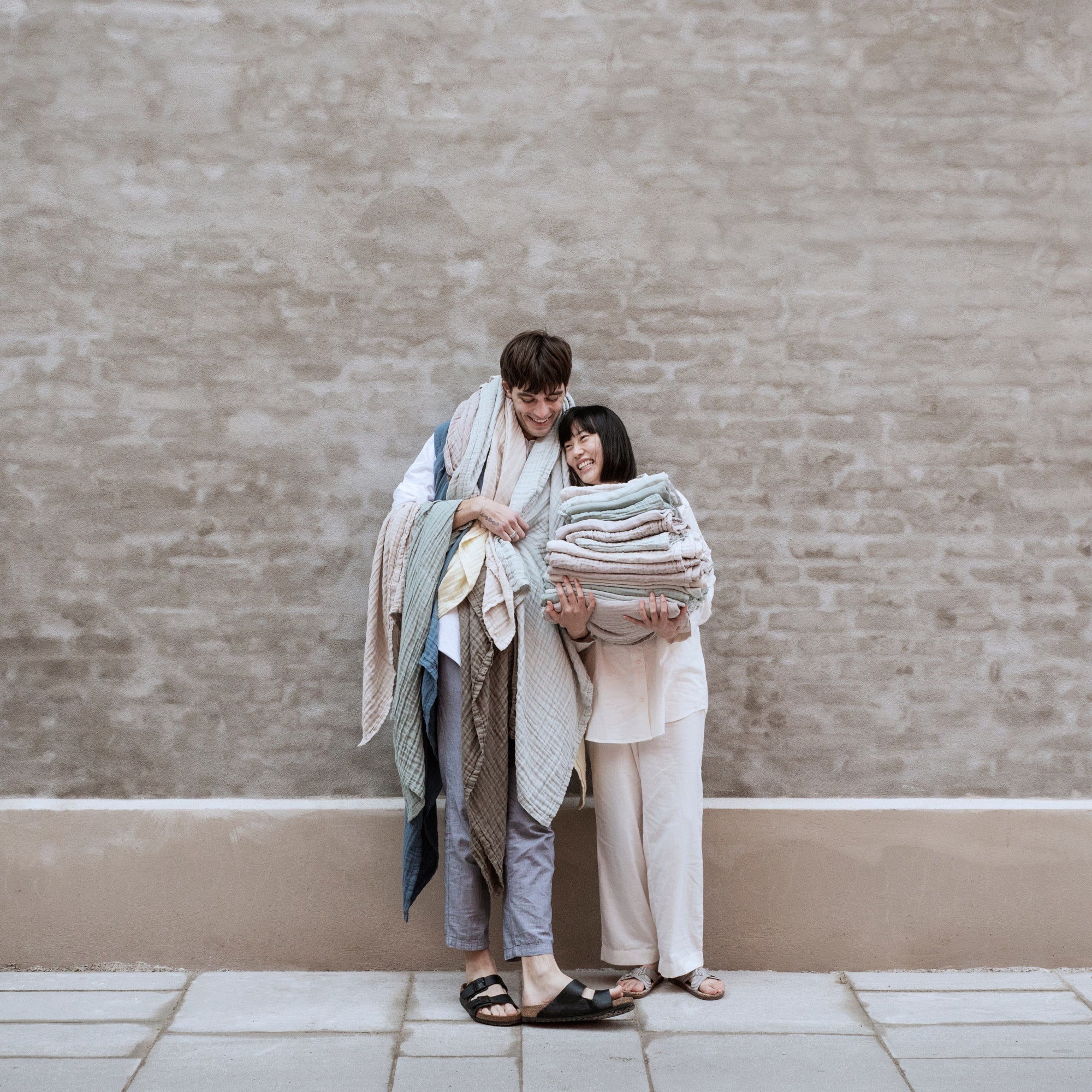 Two people holding stacks of folded organic cotton gauze bath towels against a beige brick wall.