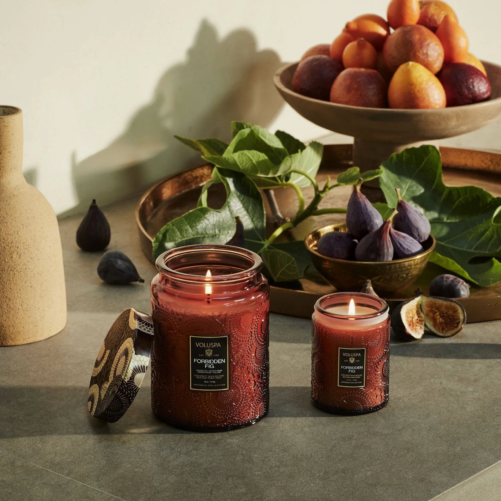 Forbidden Fig candles displayed on a table with assorted fruits including figs, nectarines, and oranges. 