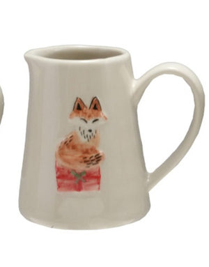An off white ceramic creamer pitcher featuring a hand-painted illustration of an orange fox sitting on a red gift box, set against a plain white background.