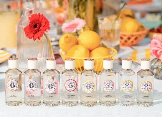 Eight small clear glass fragrance mist bottles from Roger & Gallet with white caps and round multi-colored labels are lined up on a white tablecloth. The background is a softly blurred decorative scene with lemons, flowers, and glassware.