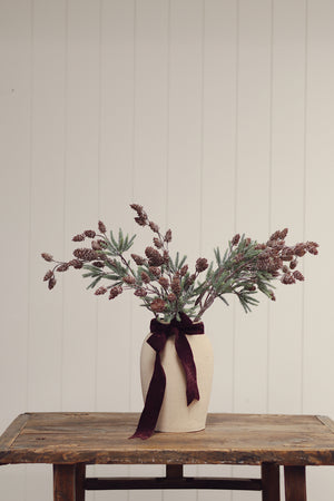 A rustic dried floral arrangement in a cream ceramic vase tied with a dark burgundy ribbon, displayed on a weathered wooden table against a light beige paneled wall.