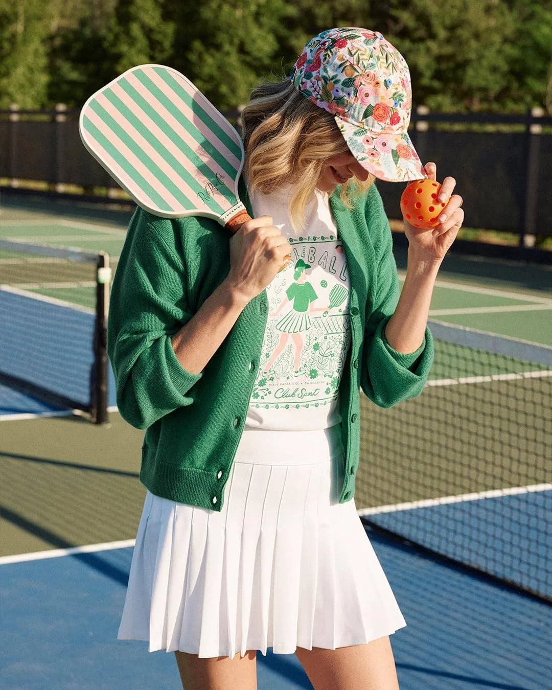 Person on a tennis court holding a paddle and ball, wearing a green cardigan, white skirt, garden party baseball cap. Rifle Paper Co. 
