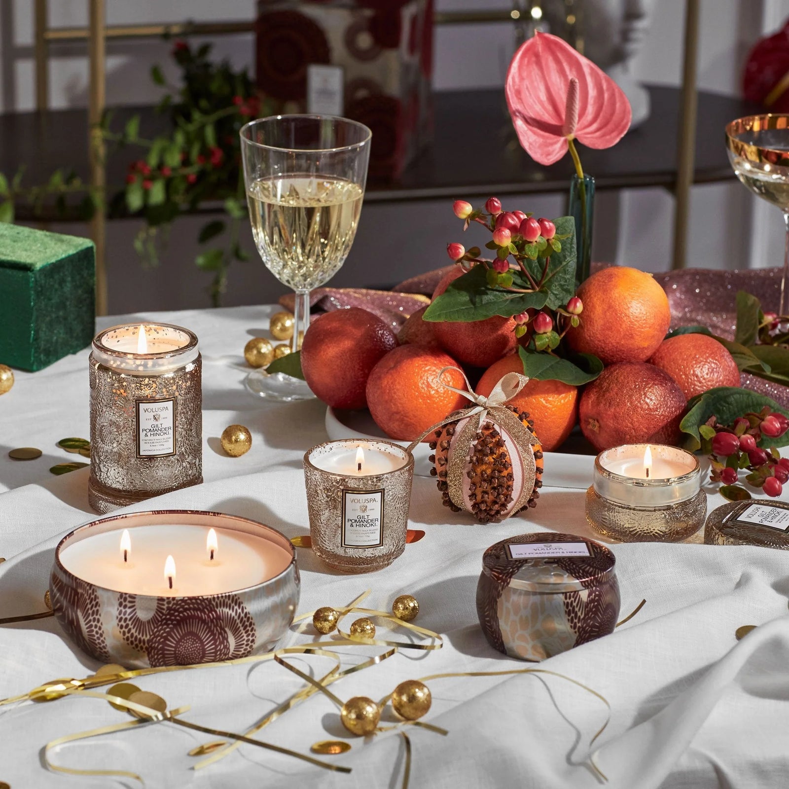 Decorative table setting with Gilt Pomander Hinoki candles, various fruits, and wine glasses on a white tablecloth. Voluspa.