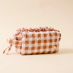 A quilted cosmetic pouch with a brown and white gingham pattern, featuring a ruffled top edge, a matching wrist strap, and a small gold button closure, set against a solid beige background.