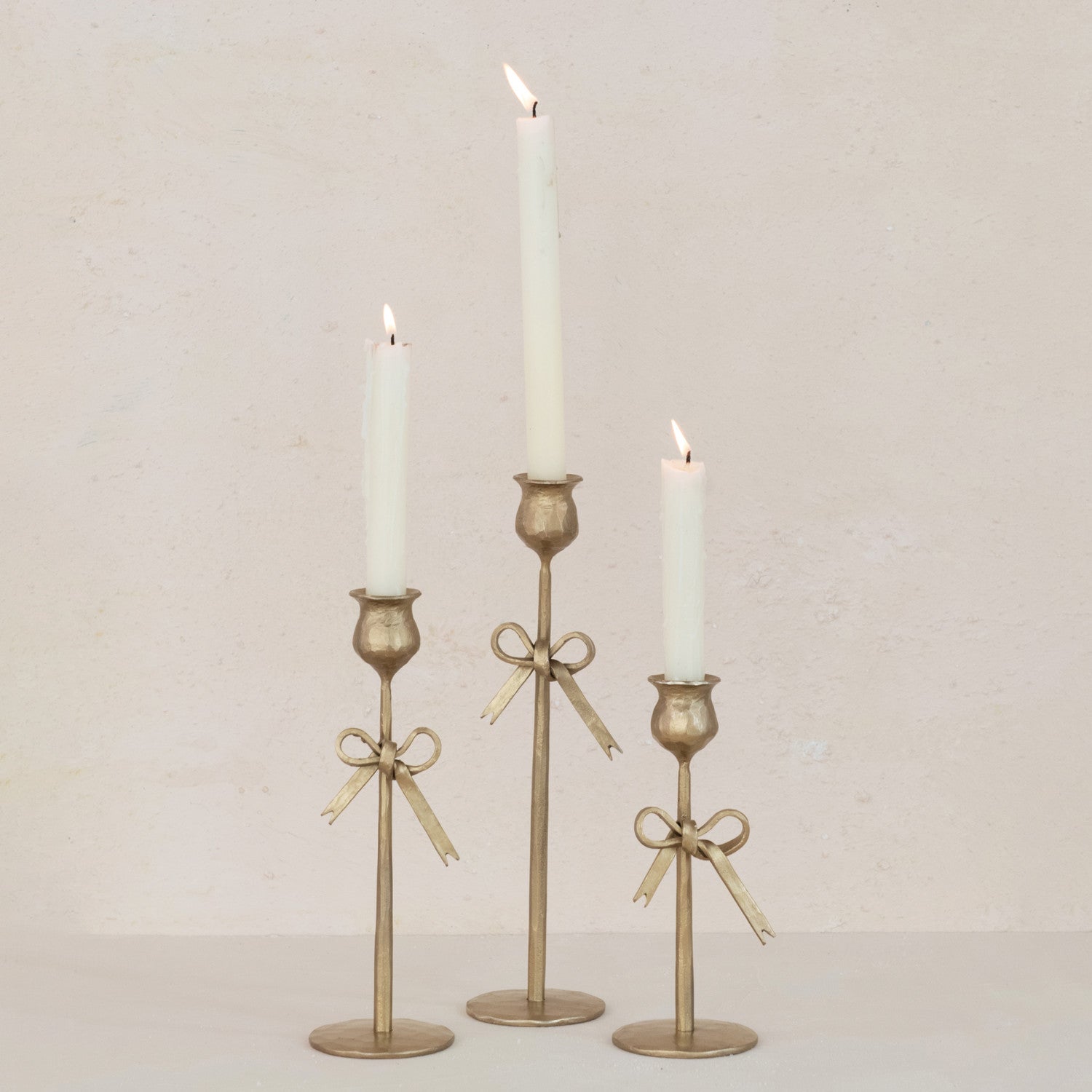 Three gold iron taper candle holders with gold bow accent detail in center against a plain background. Small, medium, and large. 