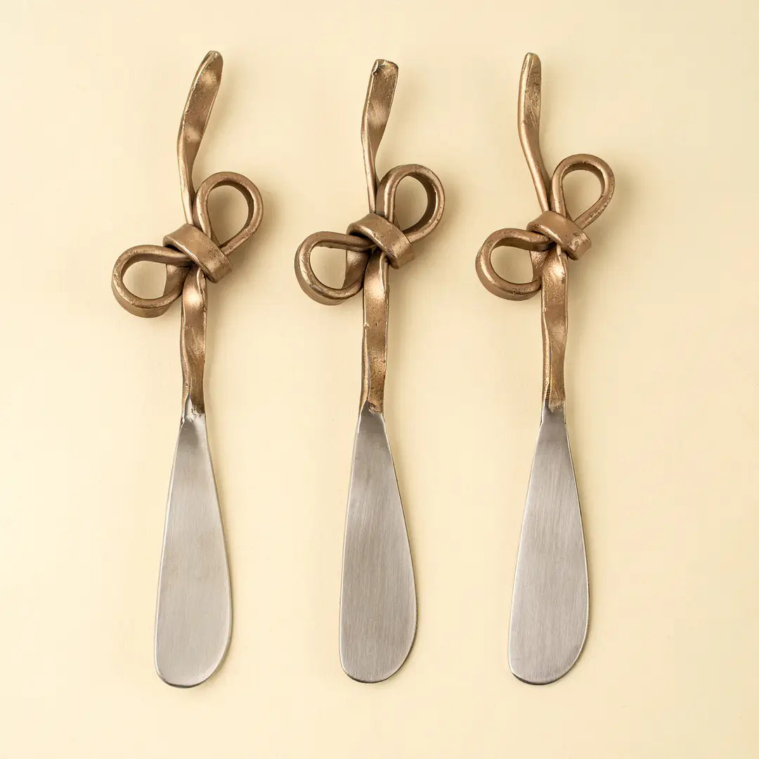 Three gold steel cheese knives with twisted bow-like handles and flat, broad blades, arranged side by side on a light cream background.