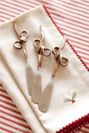 Three decorative gold steel cheese knives with knotted bow handles rest on a folded cream linen napkin with red embroidered holly, set on a red and white striped tablecloth.